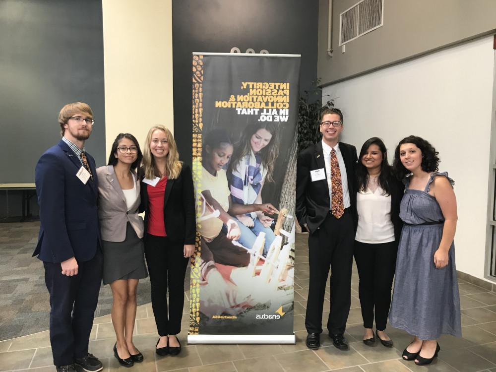 Four female and two male student members of Enactus stand in front of an Enactus banner. They are formally dressed.